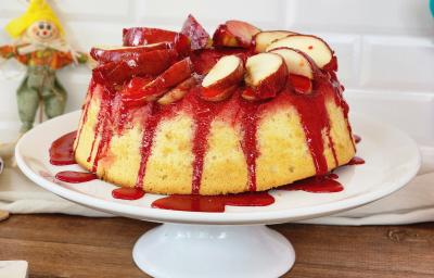 Foto da receita de bolo de maçã do amor, servido em um prato alto branco, com calda vermelha de maçã do amor escorrendo pelas laterais, com pedaços de maçã decorando o topo e itens de festa junina sobre a bancada ao lado do bolo Foto da receita de bolo de maçã do amor, servido em um prato alto branco, com calda vermelha de maçã do amor escorrendo pelas laterais, com pedaços de maçã decorando o topo e itens de festa junina sobre a bancada ao lado do bolo