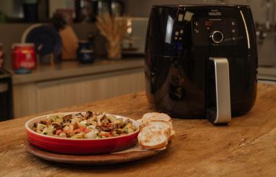Fotografia em tons de marrom de uma bancada de madeira com uma tabua de madeira, sobre ela um recipiente redondo vermelho com a caponata de legumes. Ao fundo uma airfryer.