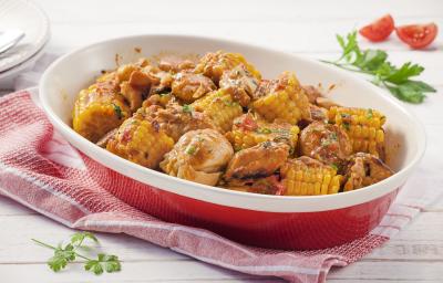 Fotografia em tons de amarelo, vermelho e branco de uma bancada de madeira branca com paninho vermelho, sobre ele um recipiente oval vermelho com bordas brancas com pedaços de frango e milho. Ao fundo duas fatias de tomate e um prato redondo branco.