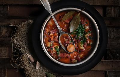 Fotografia vista de cima de uma sopa com linguiça, feijão, cenoura, costela e duas folhas de loura por cima. Apoiada na sopa, uma colher de sopa, que está dentro de um recipiente fundo de alumínio, sobre um prato preto, que está sobre uma mesa de madeira. Fotografia vista de cima de uma sopa com linguiça, feijão, cenoura, costela e duas folhas de loura por cima. Apoiada na sopa, uma colher de sopa, que está dentro de um recipiente fundo de alumínio, sobre um prato preto, que está sobre uma mesa de madeira.