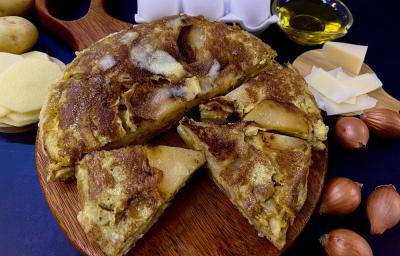 Foto em tons de amarelo da receita de tortilla de batata servida em algumas porções sobre uma tábua circular de madeira sobre uma mesa azul. Ao lado há algumas cebolas, fatias de queijo e batatas cortadas em rodelas e inteiras, além de ovos e azeite.