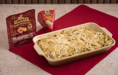 Foto da receita de Macarrão gratinado com queijo Tirolez, servida em uma travessa marrom, sobre um pano vermelho. Ao lado dela há um pacote de Queijo Parmesão Tirolez e outro de Queijo Azul Tirolez