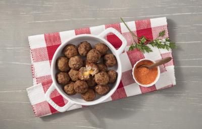 Fotografia em tons de cinza, branco e vermelho de uma bancada cinza vista de cima, ao centro um pano xadrez nas cores vermelho e branco e por cima uma panela branca e redonda com alças e dentro bolinhos de carne. Ao lado um potinho branco com molho. Fotografia em tons de cinza, branco e vermelho de uma bancada cinza vista de cima, ao centro um pano xadrez nas cores vermelho e branco e por cima uma panela branca e redonda com alças e dentro bolinhos de carne. Ao lado um potinho branco com molho.
