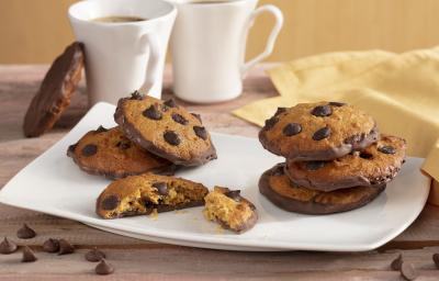Fotografia em tons de amarelo em uma bancada de madeira com um pano amarelo, um recipiente quadrado branco com alguns cookies de cenoura recheado com chocolate. Ao fundo, duas xícaras brancas com café. Fotografia em tons de amarelo em uma bancada de madeira com um pano amarelo, um recipiente quadrado branco com alguns cookies de cenoura recheado com chocolate. Ao fundo, duas xícaras brancas com café.
