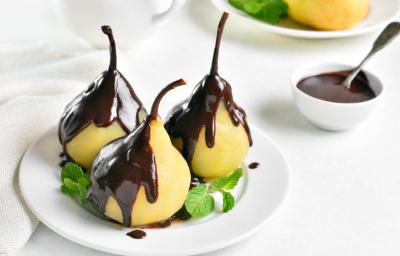 Fotografia de uma sobremesa de peras cozidas no suco de laranja com uma cobertura de chocolate. A sobremesa está dentro de um prato pequeno de vidro com pequenas folhas de hortelã, sobre uma mesa branca. Ao fundo, um pote pequeno com calda de chocolate. Fotografia de uma sobremesa de peras cozidas no suco de laranja com uma cobertura de chocolate. A sobremesa está dentro de um prato pequeno de vidro com pequenas folhas de hortelã, sobre uma mesa branca. Ao fundo, um pote pequeno com calda de chocolate.