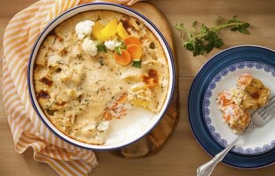 Fotografia em tons de laranja e azul em uma bancada de madeira com um pano com listras laranjas, uma tábua de madeira com um refratário grande e redondo com legumes gratinados. Ao lado, um prato branco com detalhes azuis, com um pedaço dos legumes.