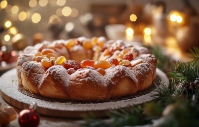 Fotografia de um bolo natalino circular, coberto com açúcar de confeiteiro e decorado com frutas secas coloridas. O fundo apresenta luzes suaves e elementos festivos.