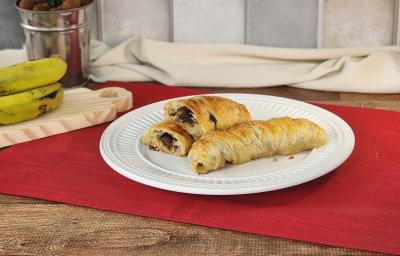 Fotografia em tons de vermelho em uma bancada de madeira escura com um pano vermelho e um prato branco redondo ao centro com duas bananas enroladas em massa folhada e recheadas de chocolate em cima. Ao fundo, cacho de banana e paninho bege.