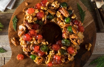 Fotografia de um bolo de frutas circular em forma de guirlanda, adornado com frutas cristalizadas e nozes, colocado sobre uma tábua de madeira rústica, cercado por ramos de pinheiro, evocando um clima natalino e aconchegante. Fotografia de um bolo de frutas circular em forma de guirlanda, adornado com frutas cristalizadas e nozes, colocado sobre uma tábua de madeira rústica, cercado por ramos de pinheiro, evocando um clima natalino e aconchegante.