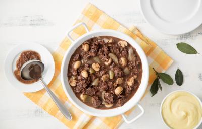 fotografia em tons de branco, amarelo e marrom vista de uma bancada branca. Contém um pano amarelo e por cima uma panela redonda branca com o Ensopado de carne com vinho. Ao lado direito um pote redondo com creme e ao lado esquerdo o ensopado é servido.