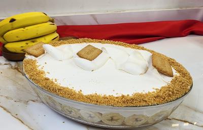Foto da receita de Torta Banoffee de Travessa, servida em uma travessa oval de vidro, decorada com chantilly batido e biscoitos tostines triturados, tudo em uma bancada de mármore com algumas bananas ao fundo e um tecido vermelho