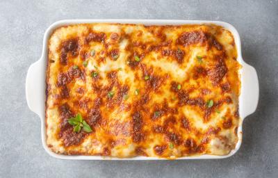 Fotografia vista de cima de uma receita com mandioca, frango e molho de tomate com um gratinado por cima de queijo dentro de uma travessa branca de vidro retangular. O recipiente está sobre uma mesa de cimento queimado.