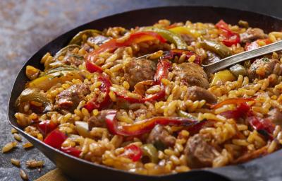 Fotografia de uma panela preta com bordas finas, com arroz amarelo, pimentão nas cores verde, amarelo e vermelho e linguiça em pedaços. No centro da panela, uma colher prata apoiada. Fotografia de uma panela preta com bordas finas, com arroz amarelo, pimentão nas cores verde, amarelo e vermelho e linguiça em pedaços. No centro da panela, uma colher prata apoiada.