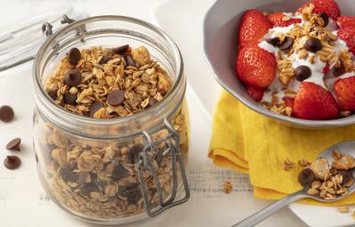 Fotografia em tons de branco de uma bancada branca com um pote de vidro com tampa e granola. Ao lado um prato branco com paninho amarelo e uma salada de frutas com granola. Fotografia em tons de branco de uma bancada branca com um pote de vidro com tampa e granola. Ao lado um prato branco com paninho amarelo e uma salada de frutas com granola.