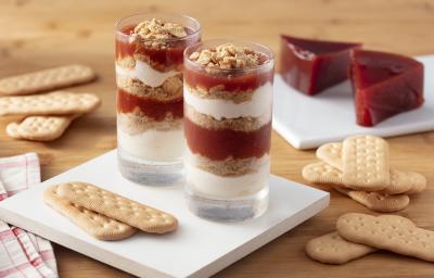 Fotografia em tons de marrom e branco de uma bancada de madeira marrom com um apoio de mesa branco quadrado, sobre ele dois copos de vidro com o verrine e biscoitos. Ao fundo biscoitos e fatias de goiabada.