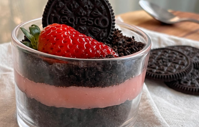 A imagem mostra um copinho de vidro, baixo e transparente, com um doce em camadas escuras e cor de rosa. Um morango e um biscoito Negresco decoram o topo. o copo está apoiado em um guardanapo de pano claro com alguns biscoitos e uma colher ao fundo.