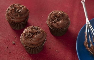 Fotografia em tons escuros de três cupcakes de nescau extra cacau com cobertura sobre um pano vermelho escuro. Ao lado tem um batedor de arame apoiado em um prato azul de vidro, raso.