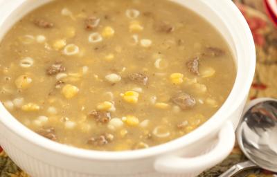 Fotografia em tons de branco e marrom de uma bancada de madeira com paninho marrom com detalhes vermelhos, sobre ele um recipiente redondo branco com a sopa de grão-de-bico. Ao lado uma colher e ao fundo dois potes redondos vermelhos. Fotografia em tons de branco e marrom de uma bancada de madeira com paninho marrom com detalhes vermelhos, sobre ele um recipiente redondo branco com a sopa de grão-de-bico. Ao lado uma colher e ao fundo dois potes redondos vermelhos.