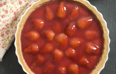 Fotografia em tons de vermelho com em uma bancada de madeira clara com um pano vermelho, um prato branco raso grande com a torta de morango. Fotografia em tons de vermelho com em uma bancada de madeira clara com um pano vermelho, um prato branco raso grande com a torta de morango.