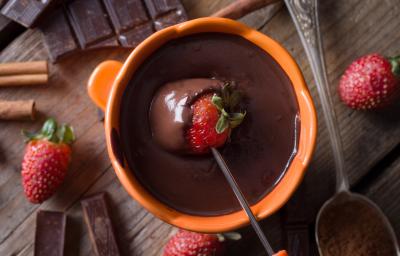 Fotografia mostra um morango mergulhado em uma panela laranja com chocolate derretido.