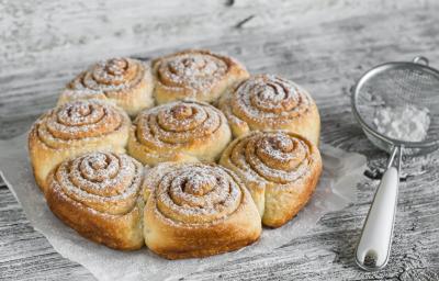 Fotografia em tons de cinza em uma bancada de madeira cinza contém diversos pães redondos com açúcar por cima, ao lado uma peneira com açúcar.