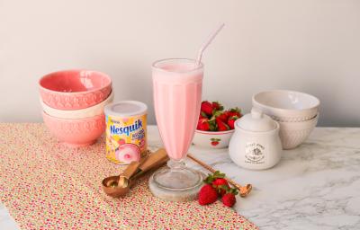 Fotografia de um milkshake rosa servido em uma copo alto de vidro. Ao fundo um cenário com morangos e bowls com tons rosas.