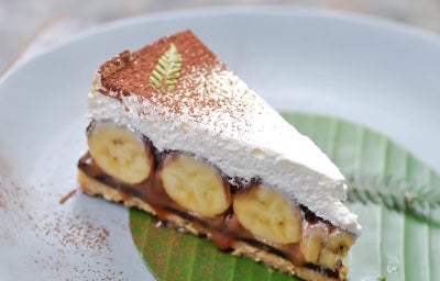 Em cima de uma mesa, prato com fatia de banoffee com massa crocante, doce de leite cremoso e bananas em rodelas, decorado com chantilly e chocolate em pó. Em cima de uma mesa, prato com fatia de banoffee com massa crocante, doce de leite cremoso e bananas em rodelas, decorado com chantilly e chocolate em pó.