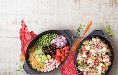 Fotografia em tons de cinza, vermelho, preto de uma bancada cinza vista de cima, contém um pano vermelho e por cima dois recipientes ovais um com macarrão, azeitona, peito de peru e tomates picados e no outro os mesmos ingredientes só que inteiros. Fotografia em tons de cinza, vermelho, preto de uma bancada cinza vista de cima, contém um pano vermelho e por cima dois recipientes ovais um com macarrão, azeitona, peito de peru e tomates picados e no outro os mesmos ingredientes só que inteiros.