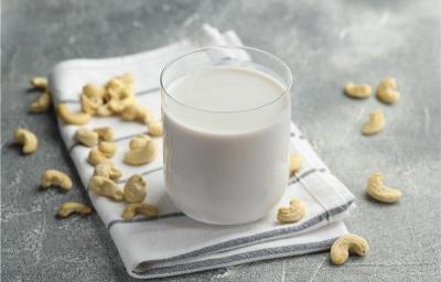 Fotografia em tons claros de um copo de vidro com uma bebida vegetal de castanha de caju apoiado em um pano branco dobrado com algumas listras escuras, que está sobre uma mesa de cimento queimado. Por cima da mesa e do pano tem castanhas espalhadas.