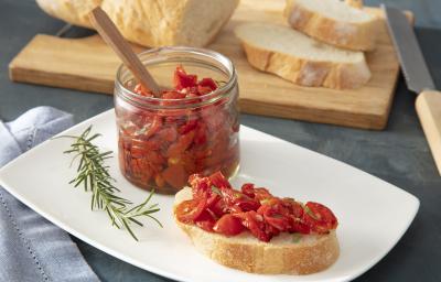 Foto da receita de Tomate confit. Observa-se uma torrada com os tomates assados em cima e um recipiente transparente de vidro com os tomates armazenados. Alecrim e pedaços de pão fatiados decoram a foto. Foto da receita de Tomate confit. Observa-se uma torrada com os tomates assados em cima e um recipiente transparente de vidro com os tomates armazenados. Alecrim e pedaços de pão fatiados decoram a foto.