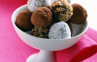 Fotografia em tom claro, um pote branco decorativo sobre uma toalha de mesa rosa. Dentro desse recipiente, tem várias trufas de chocolate, algumas enroladas em nozes trituradas, outras em cacau e três delas passadas em açúcar de confeiteiro.