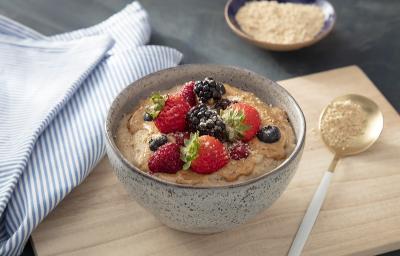 Foto em tons de bege da receita de mingau nutritivo servida em um bowl de cerâmica sobre uma tábua de madeira clara com uma colher prateada ao lado. Ao lado ainda há um paninho azul listrado e um recipiente com aveia em flocos Foto em tons de bege da receita de mingau nutritivo servida em um bowl de cerâmica sobre uma tábua de madeira clara com uma colher prateada ao lado. Ao lado ainda há um paninho azul listrado e um recipiente com aveia em flocos