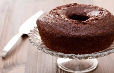 Um bolo de chocolate em forma de bundt, com cor marrom escura e superfície brilhante, está sobre um elegante suporte de vidro. O ambiente é rústico e sofisticado, sugerindo que o bolo está pronto para ser servido. Um bolo de chocolate em forma de bundt, com cor marrom escura e superfície brilhante, está sobre um elegante suporte de vidro. O ambiente é rústico e sofisticado, sugerindo que o bolo está pronto para ser servido.