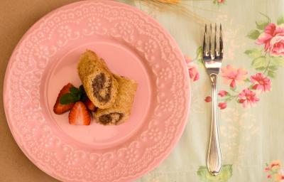 Fotografia em tons de rosa em uma mesa de madeira, uma toalha branca com flores rosas, um prato redondo raso rosa com os rolinhos recheados de chocolate e morango. Ao lado, um garfo. Fotografia em tons de rosa em uma mesa de madeira, uma toalha branca com flores rosas, um prato redondo raso rosa com os rolinhos recheados de chocolate e morango. Ao lado, um garfo.