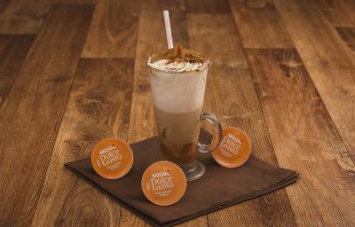 Fotografia em tons de marrom em uma bancada de madeira escura, um pano marrom, cápsulas de Mochaccino Canela Dolce Gusto e uma xícara de vidro alta com a bebida feita com banana, doce de leite e chantilly.