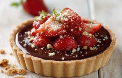 Fotografia de uma mini torta de bombom de morango, por cima tem ganahce de chocolate, alguns morangos decorando e um pouco da base triturada. A torta está sobre uma mesa de madeira clara, e ao fundo tem um morango inteiro. Fotografia de uma mini torta de bombom de morango, por cima tem ganahce de chocolate, alguns morangos decorando e um pouco da base triturada. A torta está sobre uma mesa de madeira clara, e ao fundo tem um morango inteiro.