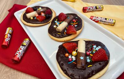 Fotografia em tons de vermelho e amarelo com um recipiente branco ao centro. Em cima do recipiente existe 3 mini pizzas doces cobertas com ganache de chocolate meio amargo, morangos e BATON Duo. Ao lado existe algumas embalagens de BATON Duo