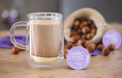 Fotografia em tons de lilás em uma bancada de madeira, uma xícara de vidro com a bebida de Mochaccino Avelã dentro, ao lado uma cápsula de Dolce Gusto, atrás um saco de juta com avelã e um paninho lilás.