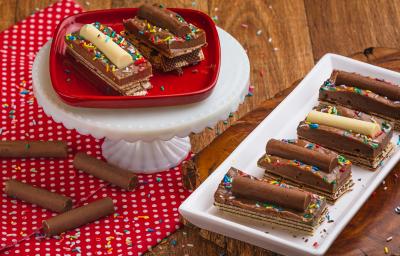 Foto de uma bancada de madeira, sobre ela há um pano vermelho com bolinhas brancas, um prato retangular com a receita pronta, alguns chocolates Baton soltos e mais um prato vermelho elevado. Há alguns granulados coloridos decorando a cena. Foto de uma bancada de madeira, sobre ela há um pano vermelho com bolinhas brancas, um prato retangular com a receita pronta, alguns chocolates Baton soltos e mais um prato vermelho elevado. Há alguns granulados coloridos decorando a cena.