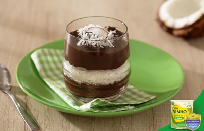 Tacinha de sobremesa em copo de vidro com camadas de chocolate e creme branco, finalizada com coco ralado, sobre prato verde.