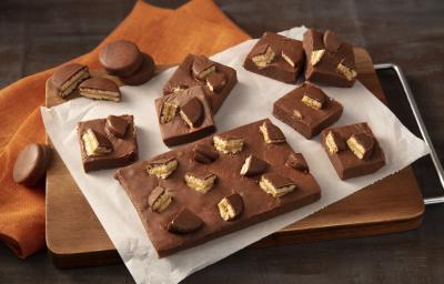 Fotografia em tons de preto e laranja de uma bancada preta com um paninho laranja, sobre ele uma tabua de madeira com um papel manteiga e o fudge de bono doce de leite. Ao lado pedaços do fudge e biscoitos bono coberto espalhados. Fotografia em tons de preto e laranja de uma bancada preta com um paninho laranja, sobre ele uma tabua de madeira com um papel manteiga e o fudge de bono doce de leite. Ao lado pedaços do fudge e biscoitos bono coberto espalhados.