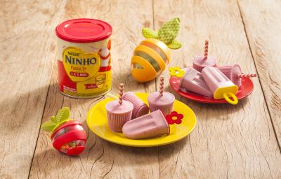 Fotografia em tons de amarelo e vermelho em uma bancada de madeira clara, dois pratos vermelho e amarelo pequenos com picolés pequenos de banana e morango. Ao lado, um lata de Ninho Fases e brinquedinhos infantis.