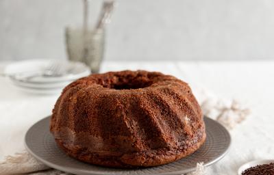 Fotografia de um bolo de chocolate feito em forma com furo central sobre um prato raso de vidro cinza. Ao lado tem granulado de chocolate em um prato pequeno raso. No fundo tem pratos brancos e garfo, e o bolo está sobre um pano branco. Fotografia de um bolo de chocolate feito em forma com furo central sobre um prato raso de vidro cinza. Ao lado tem granulado de chocolate em um prato pequeno raso. No fundo tem pratos brancos e garfo, e o bolo está sobre um pano branco.