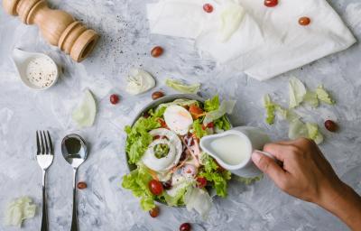 Fotografia vista de cima de uma mão segurando uma jarra pequena na cor branca, que está caindo um molho branco em uma salada de folhas, cebola, ovo cozido, maçã, tomates cereja, cenoura e azeitona preta. O refratário está sobre uma mesa de mármore branca. Fotografia vista de cima de uma mão segurando uma jarra pequena na cor branca, que está caindo um molho branco em uma salada de folhas, cebola, ovo cozido, maçã, tomates cereja, cenoura e azeitona preta. O refratário está sobre uma mesa de mármore branca.