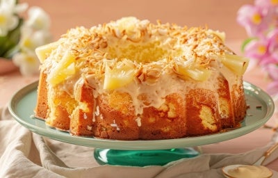 Um bolo de abacaxi com coco em formato de anel, coberto com uma calda branca cremosa, pedaços de abacaxi e coco tostado por cima. O bolo está sobre um prato verde claro e é decorado com flores ao fundo, em um cenário claro e delicado.