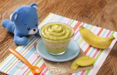 Fotografia em tons de verde em uma bancada de madeira, com um paninho listrado colorido e uma tacinha transparente com abacate e banana apoiado em um pratinho azul. Ao lado, um ursinho de pelúcia azul, uma colher laranja e uma banana.
