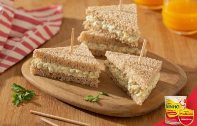 Sanduiches triangulares de pão integral recheados com ovos cremosos, presos com palitos, sobre tábua de madeira.
