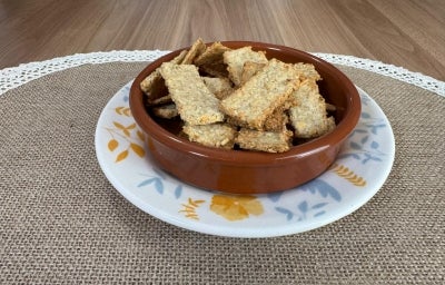 Fotografia de um prato de vidro raso branco com cores claras, sobre ele tem um recipiente pequeno fundo de tom marrom e redondo com vários biscoitinhos de aveia com queijo. Fotografia de um prato de vidro raso branco com cores claras, sobre ele tem um recipiente pequeno fundo de tom marrom e redondo com vários biscoitinhos de aveia com queijo.