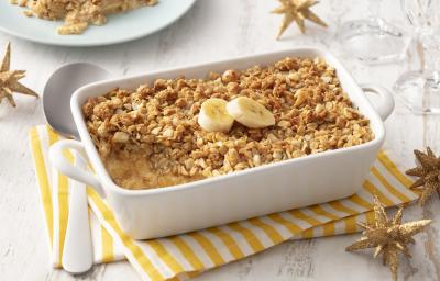 Fotografia em tons de branco e amarelo de uma bancada branca com um paninho listrado amarelo e branco, sobre ele um recipiente retangular branco com o crumble e uma colher. Ao fundo um prato com um pedaço de crumble. Fotografia em tons de branco e amarelo de uma bancada branca com um paninho listrado amarelo e branco, sobre ele um recipiente retangular branco com o crumble e uma colher. Ao fundo um prato com um pedaço de crumble.