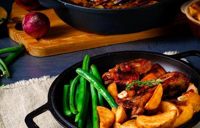 Fotografia de alguns pedaços de frango assado com batatas, vagens e ervilha torta dentro de uma panela rasa de ferro na cor preta, que está em cima de um pano branco. Ao fundo, outra panela de ferro preta com o resto dos frangos assados sobre uma tábua.